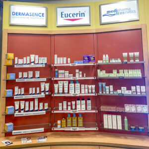 Ein Wandregal mit Kosmetikartikeln. Darüber hängen Schilder mit den Logos der Marken Dermasence, Eucerin und medipharma cosmetics