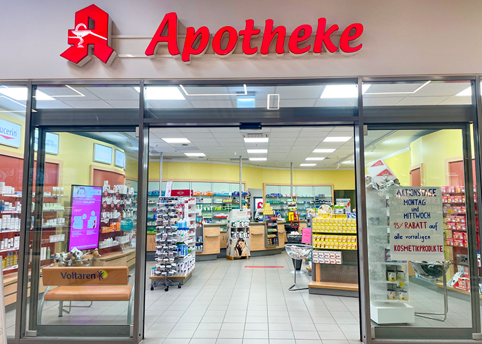 Außenansicht der Apotheke am Margaretenhof. An der Wand hängt eine rote Leuchtreklame mit dem Apotheken-A und "Apotheke", darunter sieht man die offene Tür und den Verkaufsraum dahinter